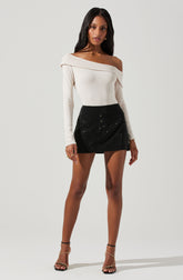 A woman is wearing a white, off-the-shoulder top with long sleeves, featuring a relaxed fit and a wide neckline that drapes over one shoulder. She pairs it with a high-waisted, black mini skirt that has a subtle sparkle or sequin embellishment. She completes the look with strappy high-heeled sandals.